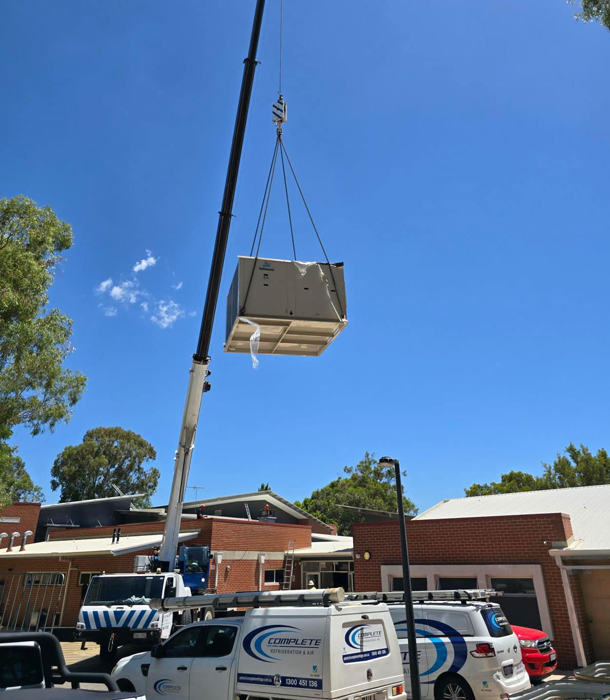 Crane lifting a large HVAC unit onto a brick building under a clear blue sky.