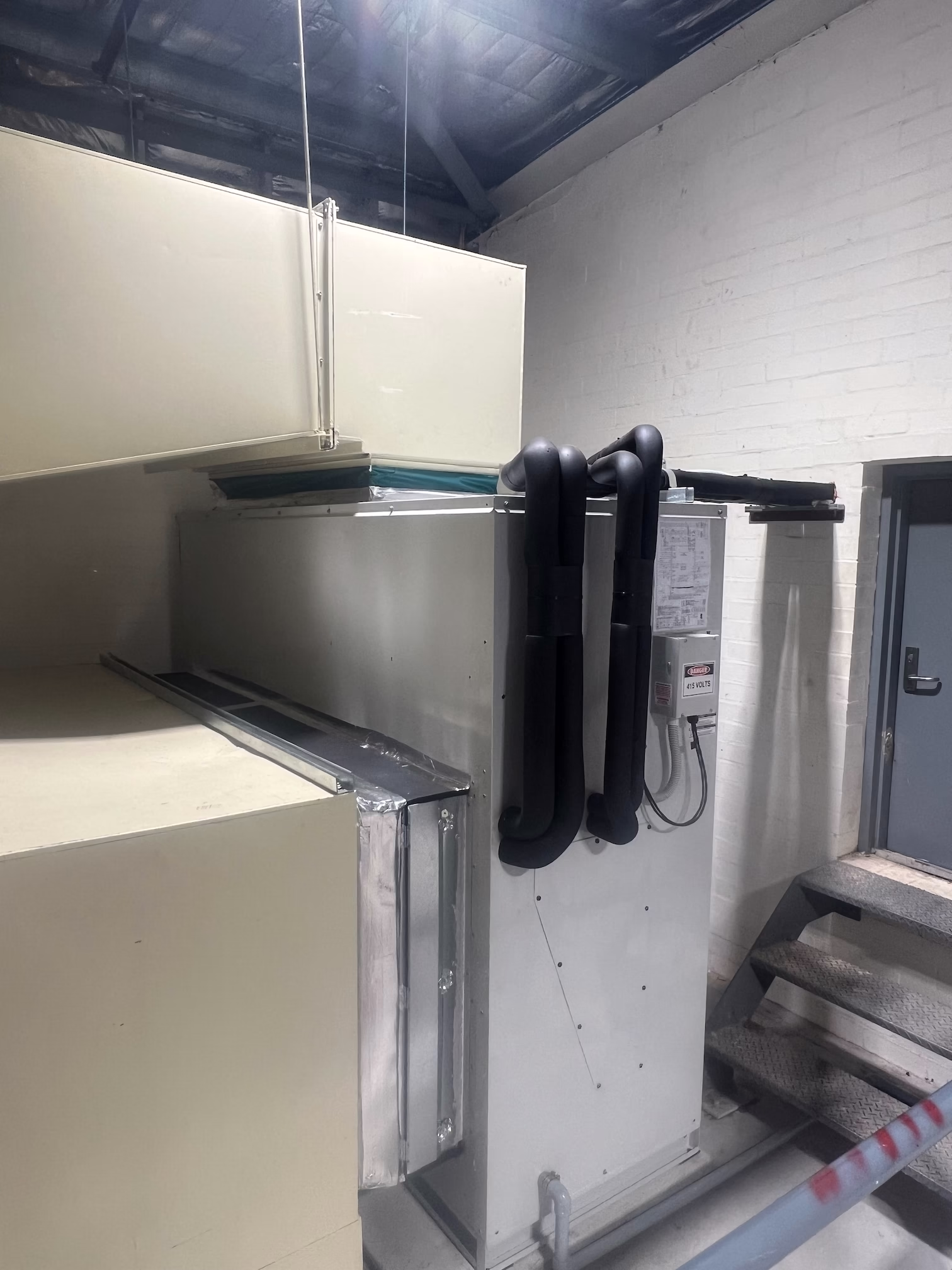 Indoor air handling unit connected to insulated pipework in plant room beside metal stairs.