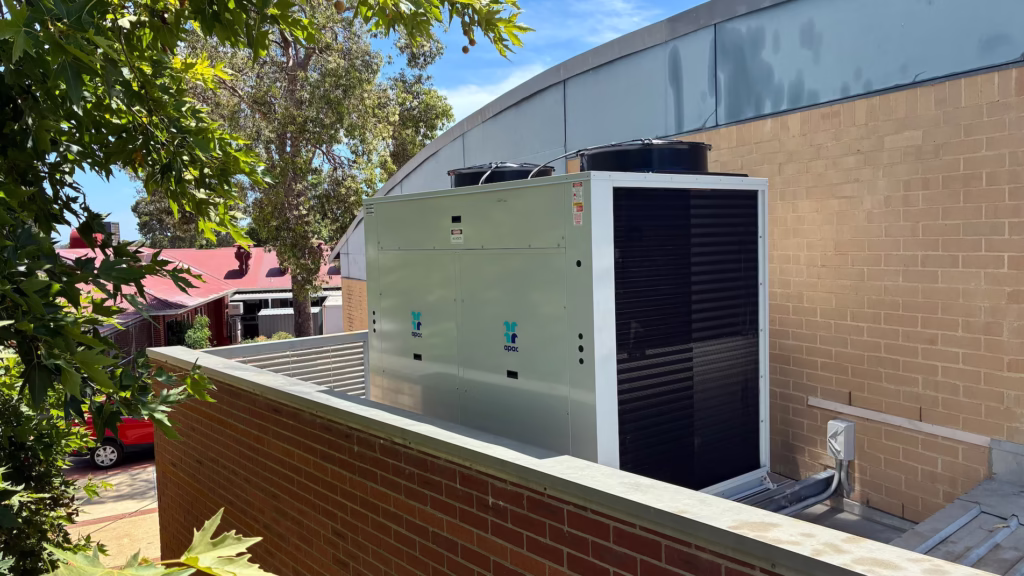 Newly installed rooftop commercial HVAC condenser unit beside brick wall with trees in background.