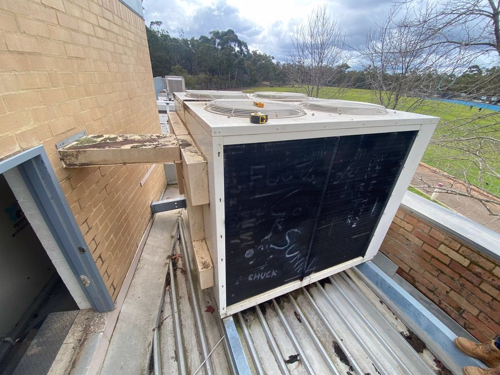 Existing rooftop HVAC condenser unit mounted on external platform beside brick building wall.