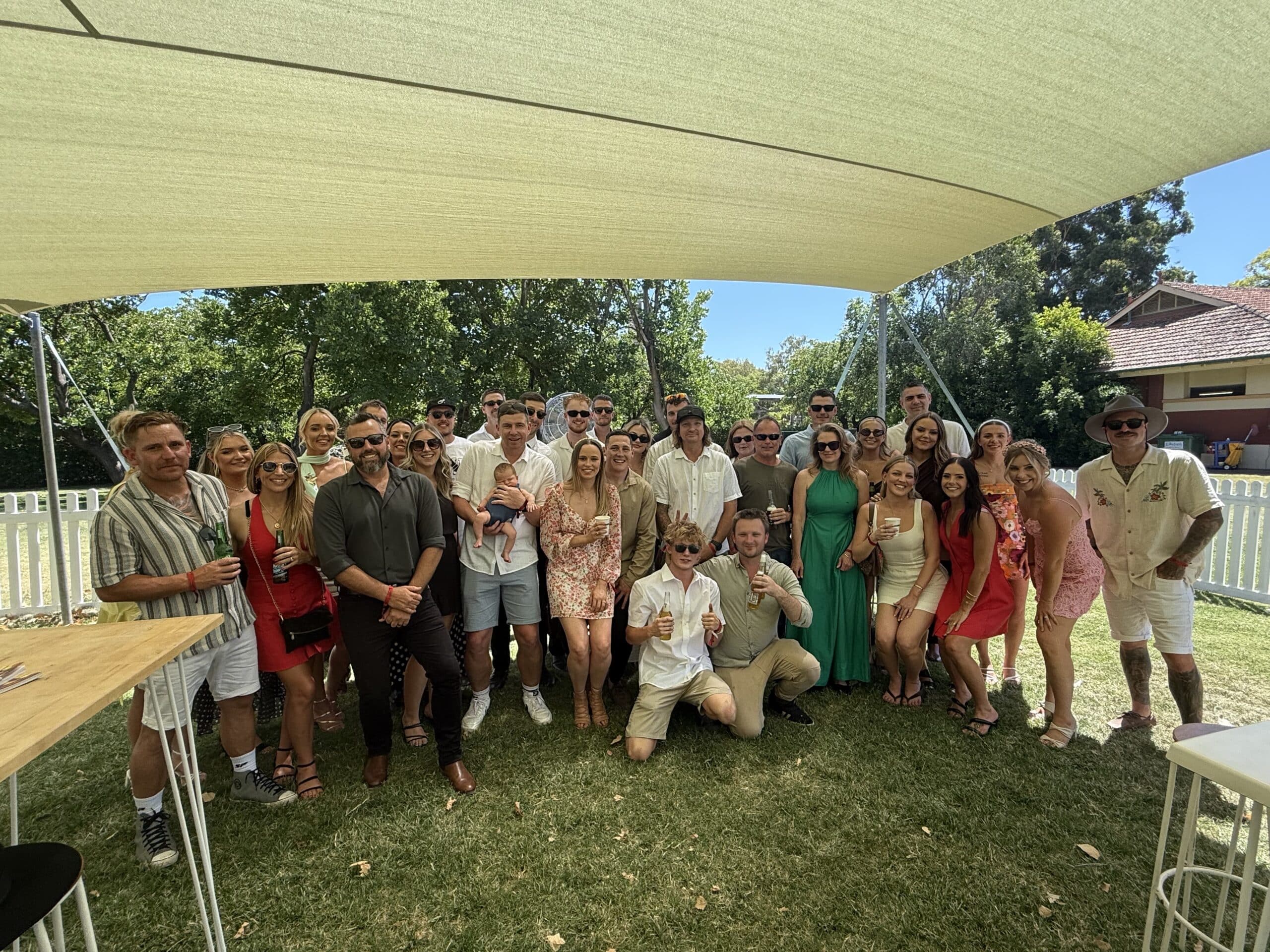 Group of guests posing together under a shade sail at an outdoor garden event.