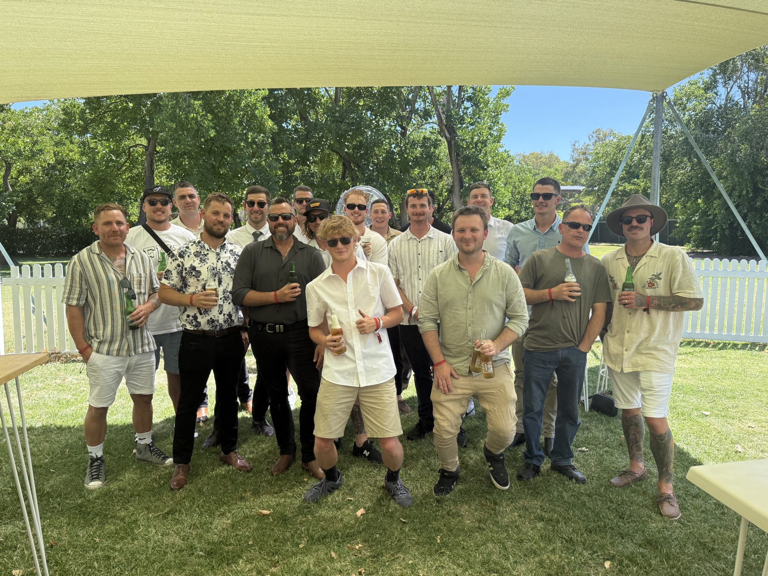Group of men standing together with drinks under a shade sail at an outdoor gathering.